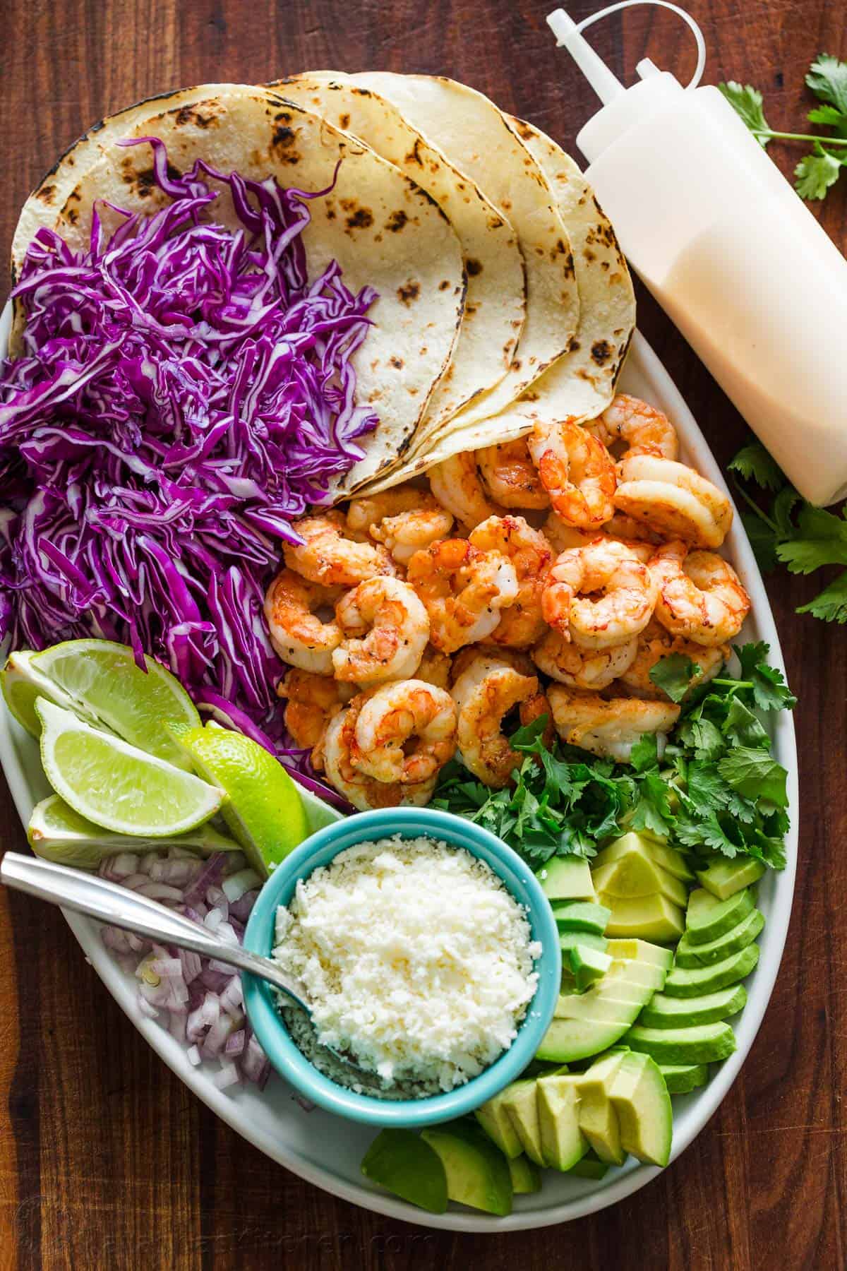 White serving platter for assembling Mexican seafood dinner cabbage, lime wedges, tortillas, onion, cotija cheese, avocado, and cilantro