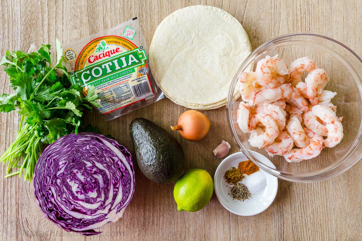 Shrimp Taco Ingredients with corn tortillas, raw shrimp, seasonings in bowl, avocado, cabbage and cotija cheese