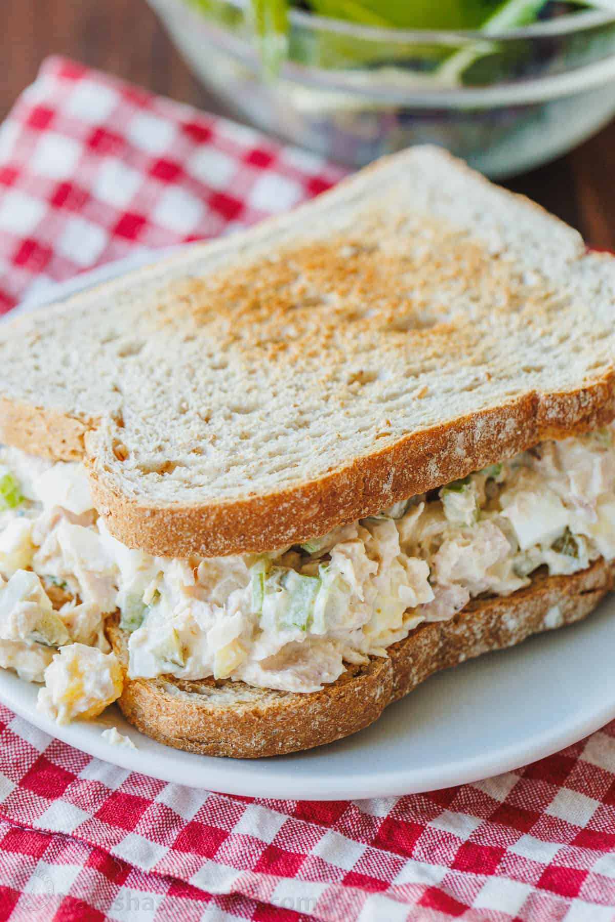 This Classic Tuna Salad recipe is light and fresh, made with tuna, celery, eggs, and the best tuna salad dressing. Great for meal prep lunch! A classic tuna salad recipe served on a sandwich on a white plate, with a red and white checked cloth underneath.